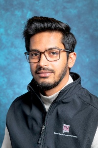 An image of Sarhan Osman Bhuiya, the Graduate Assistant for EID. The image is a head-and-shoulders portrait of a Sarhan against a textured blue background. The person has short, dark hair with a slight wave and a well-groomed beard and mustache. He is wearing rectangular glasses with brown frames and are gazing directly at the camera with a neutral expression. He is dressed in a black zip-up vest over a light gray turtleneck. The vest has an emblem on the left chest area with the text "NM STATE" in maroon and a small patch of additional text in white below it.